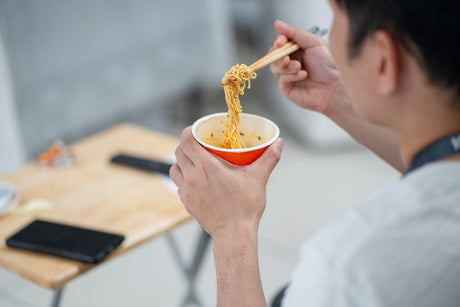 אדם אוכל אטריות מכוס נייר כתומה, האטריות מורמות במקלות אכילה מעל הכוס