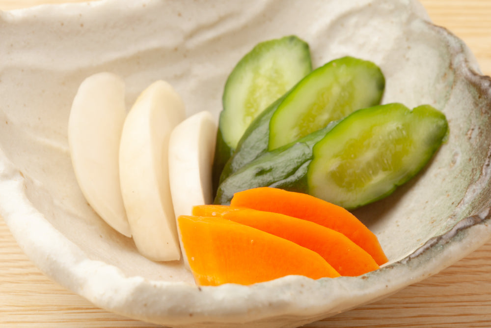 קערת קרמיקה בהירה ומחוספסת עם פרוסות מלפפון ירוקות, פרוסות גזר כתומות ופרוסות לבנות עבות, על שולחן עץ בהיר